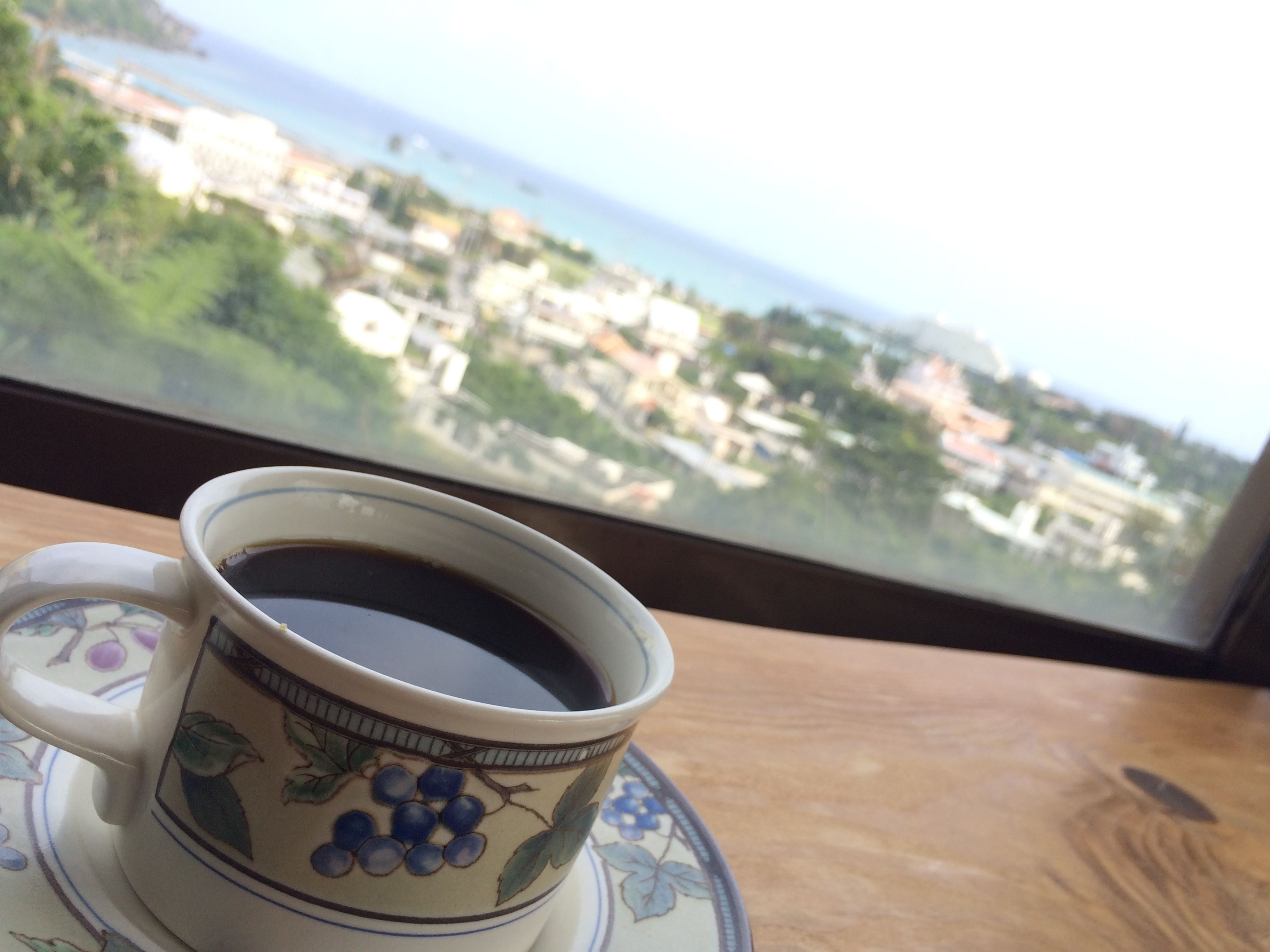 A cup of coffee on a saucer with a floral design, placed on a table by a window with a view of a cityscape.
