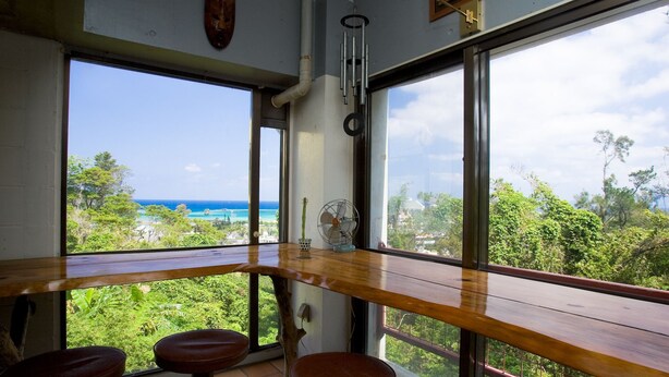 A room with a large window offering a view of the ocean and greenery. There is a wooden table with two stools and a fan. - Surfside Bed & Breakfast (Onna)