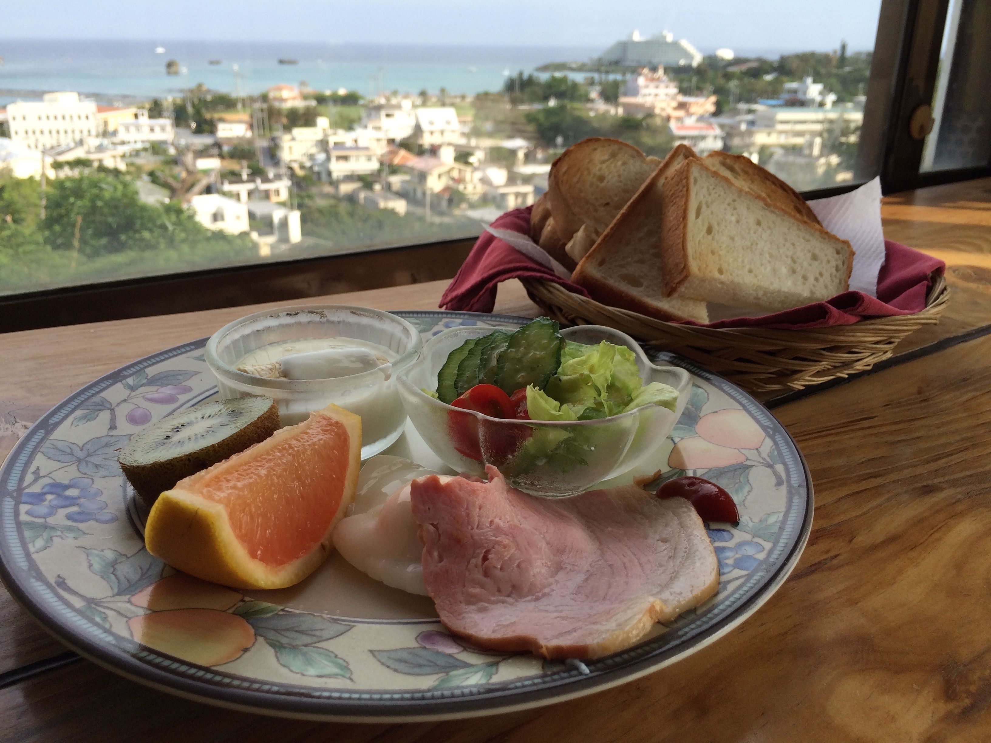 A plate with food items including ham, a slice of orange, and a salad, with a view of buildings and the sea in the background.