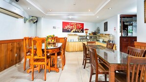 Un espace repas avec des chaises en bois et une table en verre, un buffet garni de plats et de boissons, et un ventilateur fixé au mur.