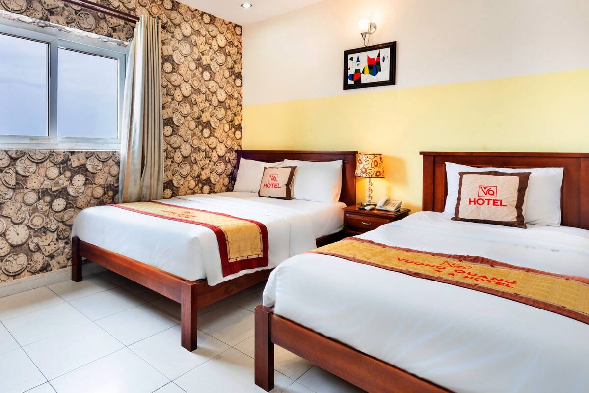 A hotel room with two beds, a window, a clock-themed wallpaper, a wall-mounted picture, and a bedside table with a lamp.
