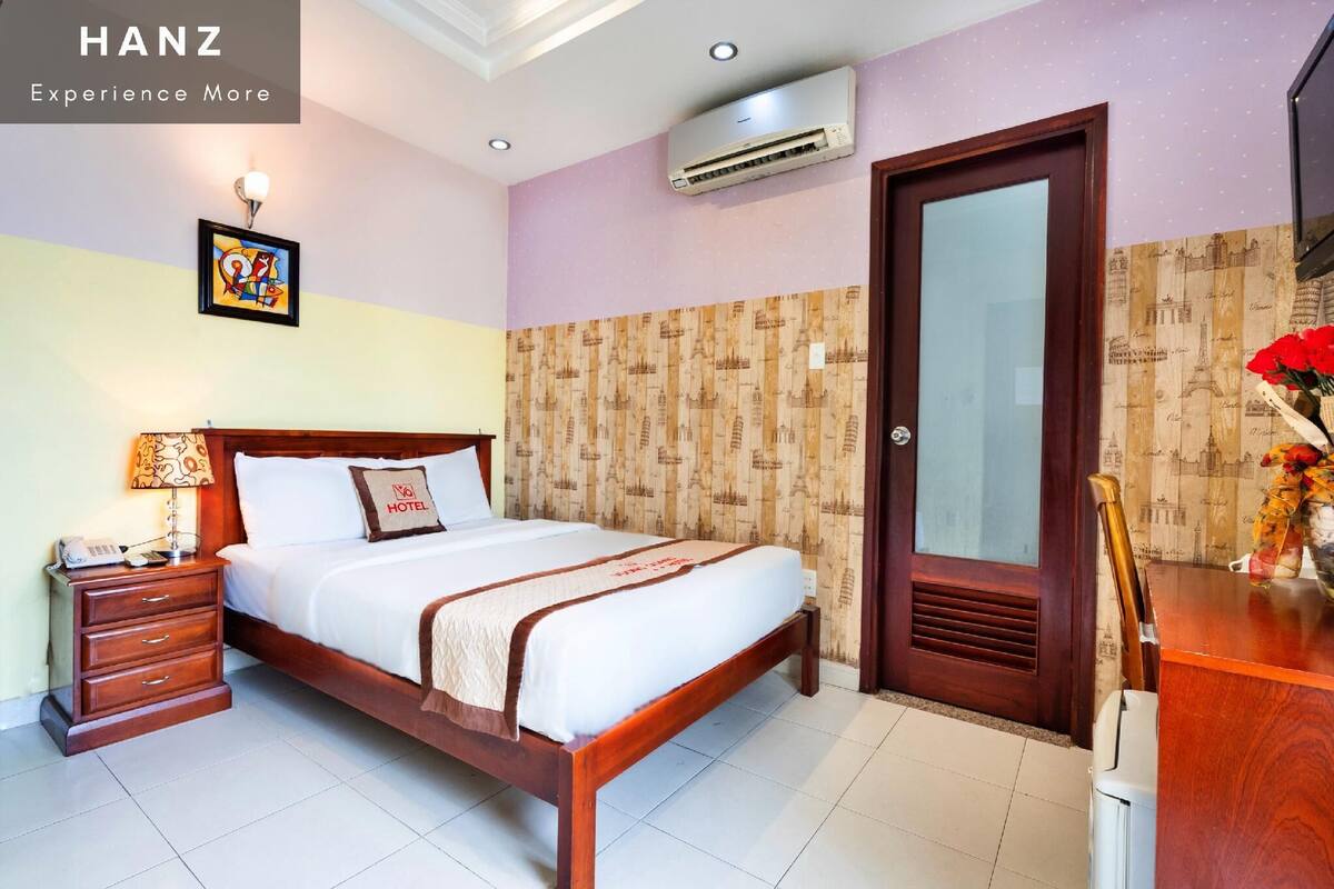 A hotel room with a bed, bedside table, lamp, air conditioning unit, and a wooden door.