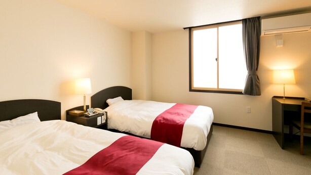 Una habitación de hotel con dos camas, cada una con una colcha roja y blanca, una mesita de noche con una lámpara y una ventana con cortinas.