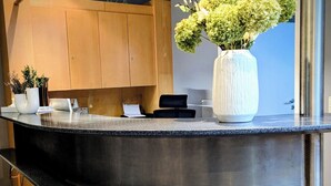 A reception desk with a curved design, a large white vase with green plants, wooden cabinets, and a metal pillar. - Hotel Herian (Vaterstetten)
