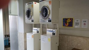 Een wasserette met drie gestapelde wasmachines, elk voorzien van een nummer. Er hangen borden aan de muur, waaronder een met een kleurrijke illustratie en tekst.