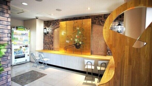 A reception area with a brick wall, a wooden counter, and a large decorative panel. There are two chairs and a vending machine in the background. - BK Weekly Mansion & Hotel (WAKAYAMA SHI)