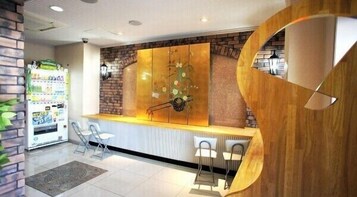 Un área de recepción con pared de ladrillo, mostrador de madera y un panel decorativo grande. Se observan dos sillas y una máquina expendedora al fondo.