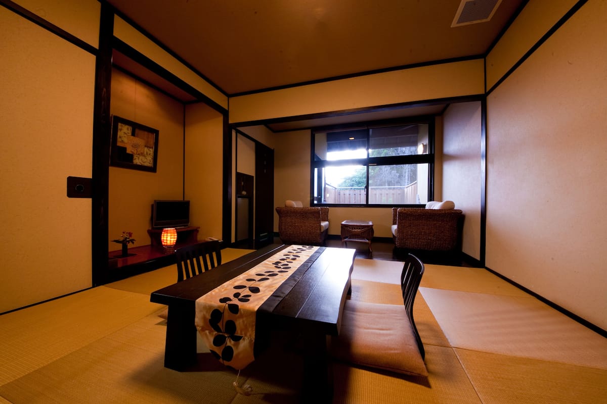 A traditional Japanese room with tatami flooring, a low table with a patterned tablecloth, a television, and a window with a view of the outside.