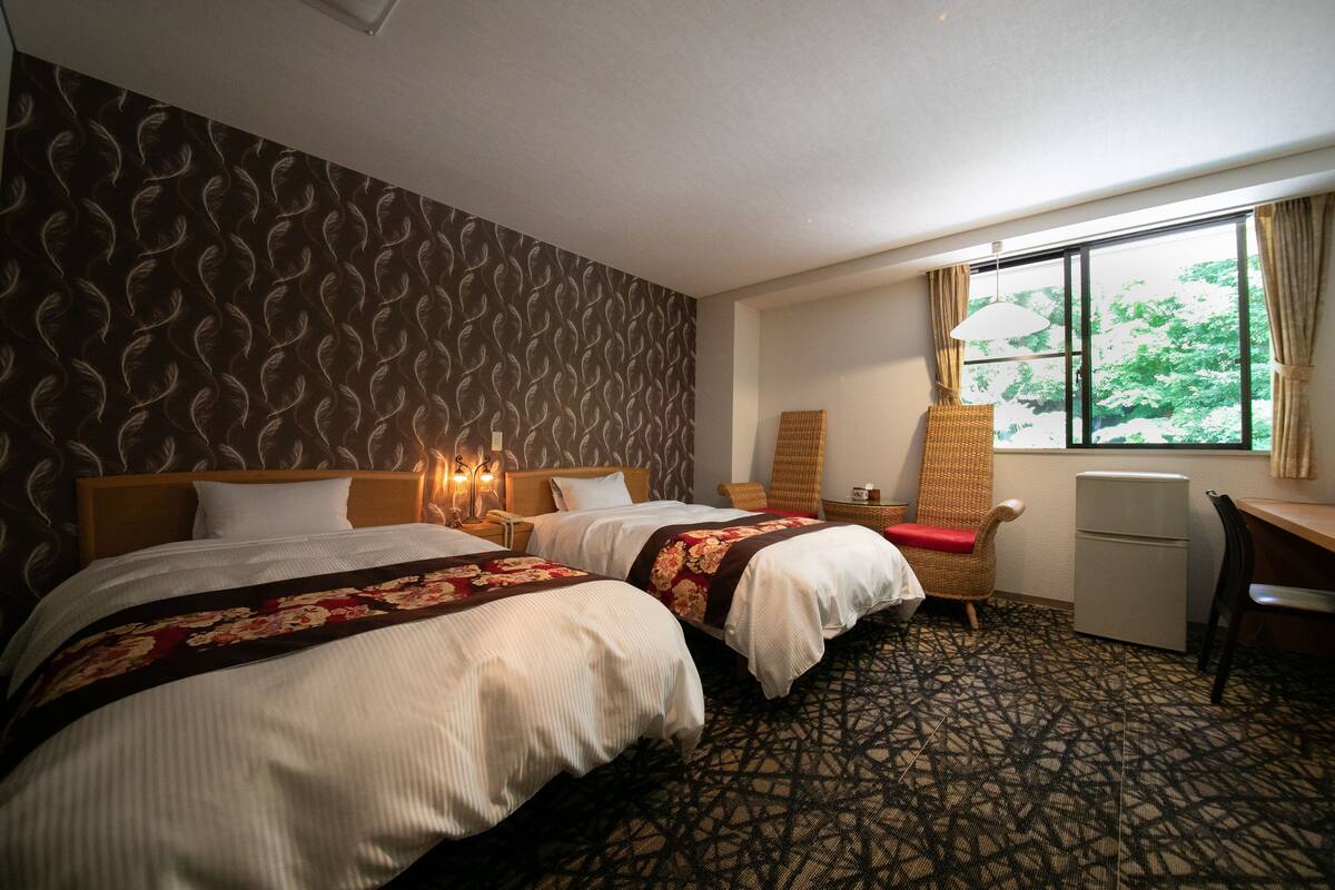 A hotel room with two beds, a desk, a chair, a window with curtains, and a patterned carpet.