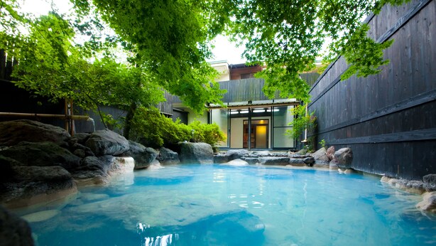 Un onsen japonés tradicional con una piscina cristalina, rodeado de formaciones rocosas naturales y exuberante vegetación.