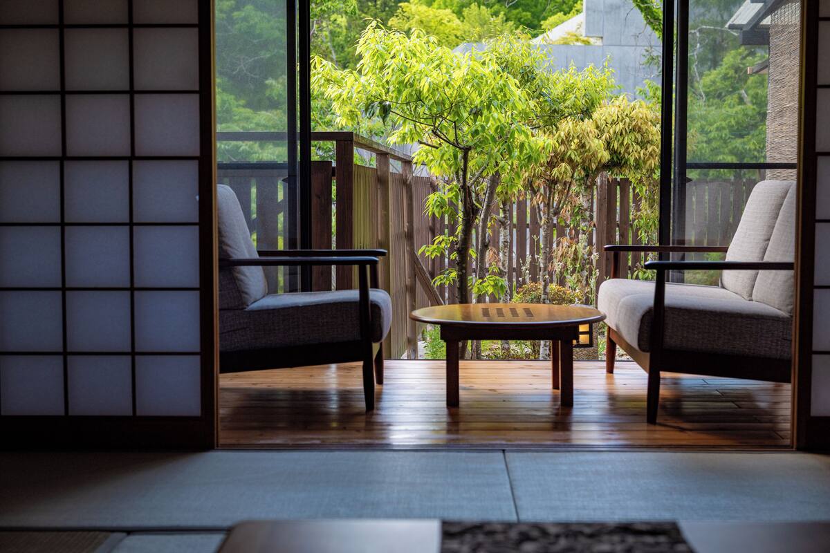 A room with tatami flooring, a low table, two chairs, and a view of a garden through large windows.