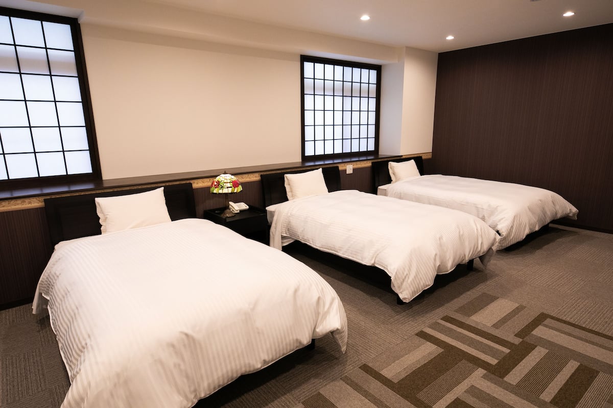 A hotel room with three single beds, a nightstand with a lamp, and a large window with a grid pattern.