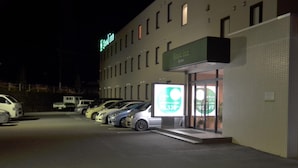 Et hotel med et skilt der viser "Good Inn Hayato" og en stor, oplyst reklame. Flere biler er parkeret foran.