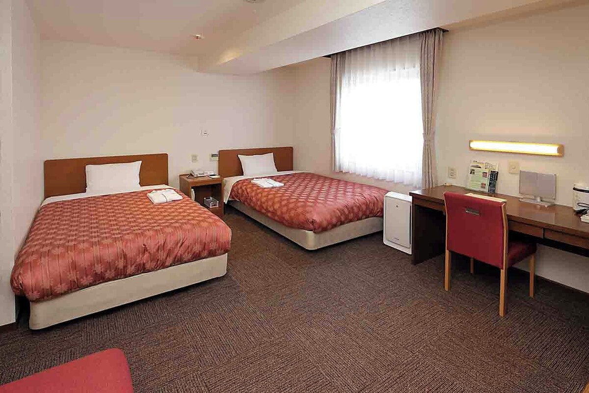 A hotel room with two beds, a desk, a chair, and a small refrigerator.