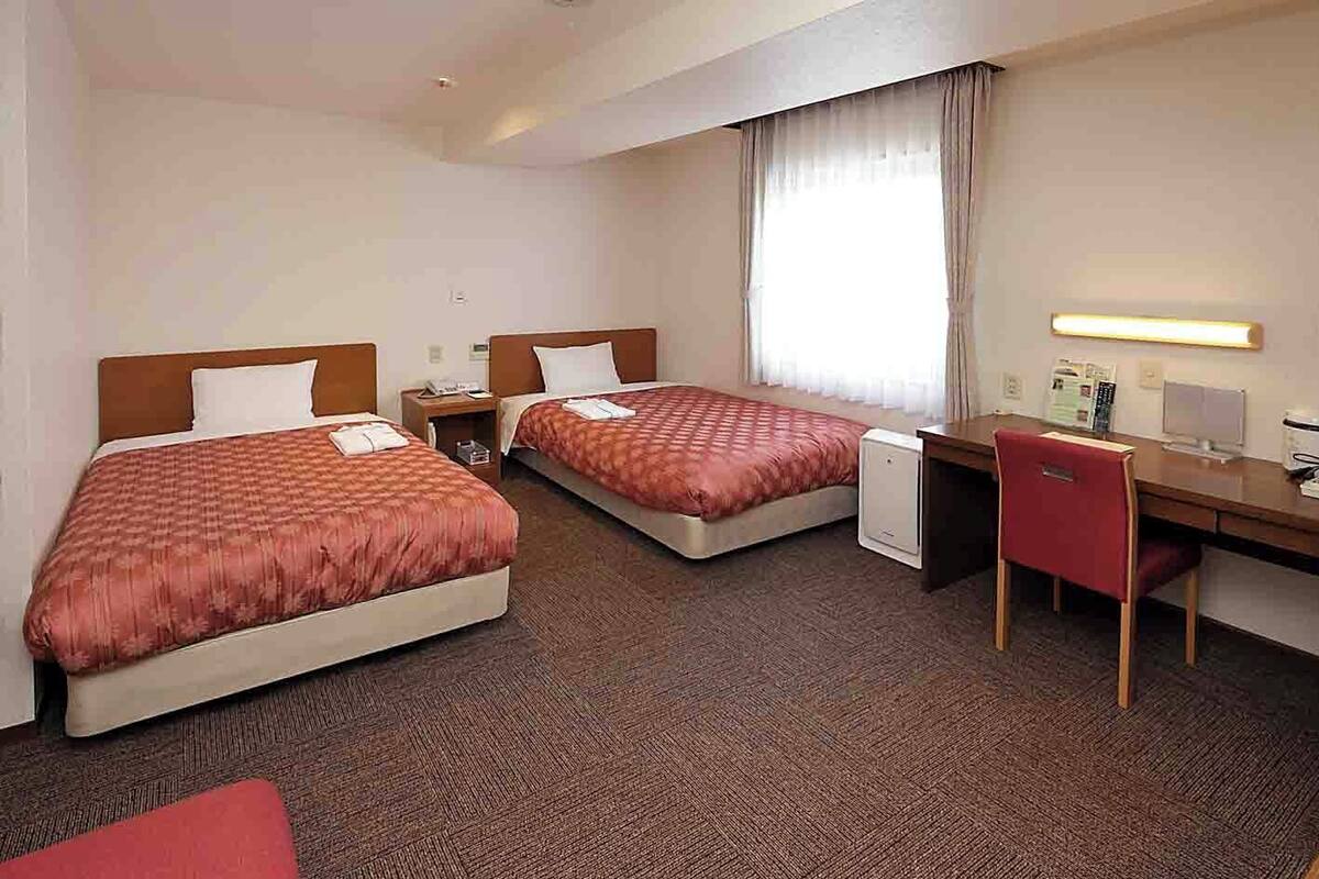 A hotel room with two beds, a desk, a chair, and a small refrigerator.