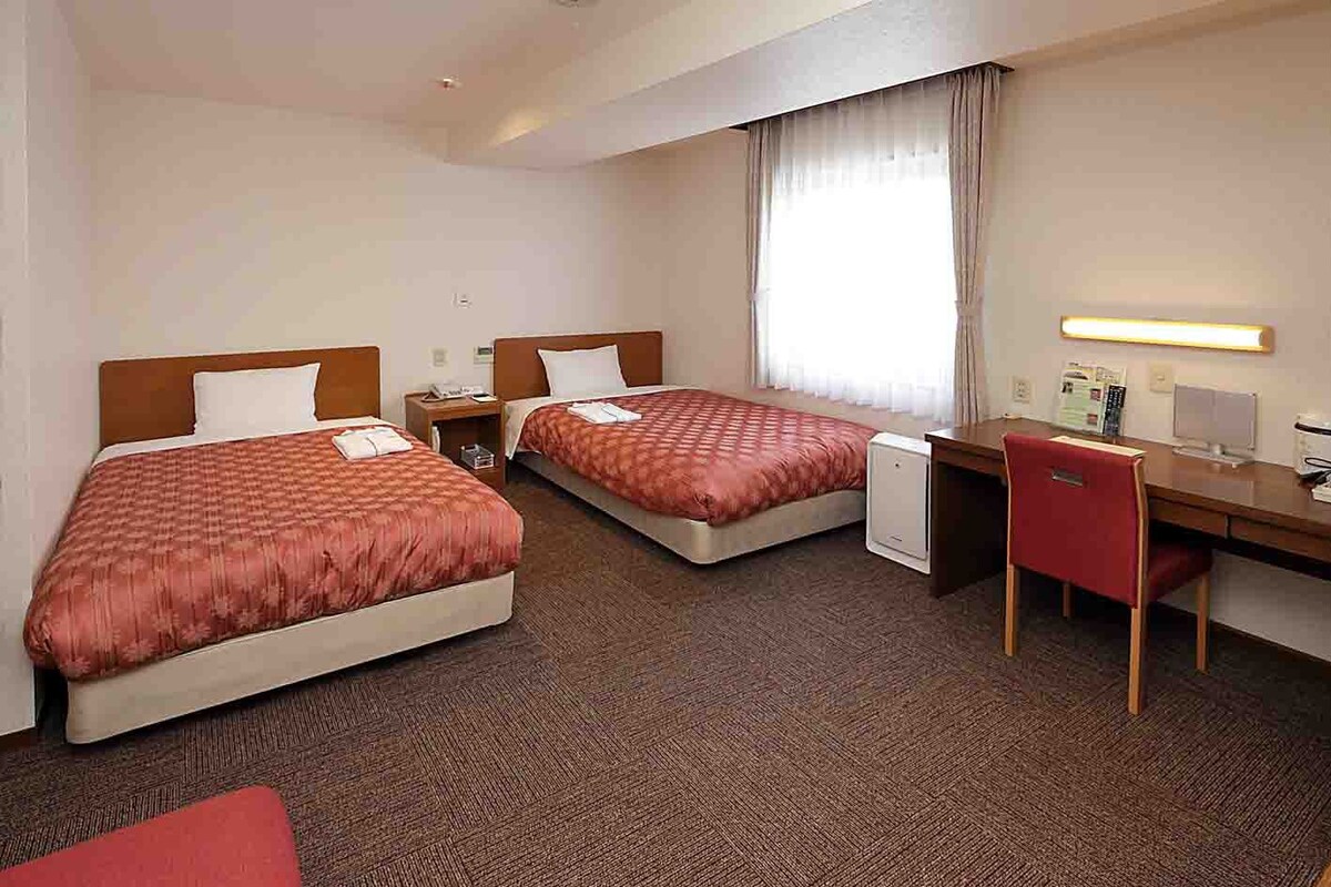 Habitación de hotel con dos camas, un escritorio, una silla, una mesita y una ventana con cortinas.