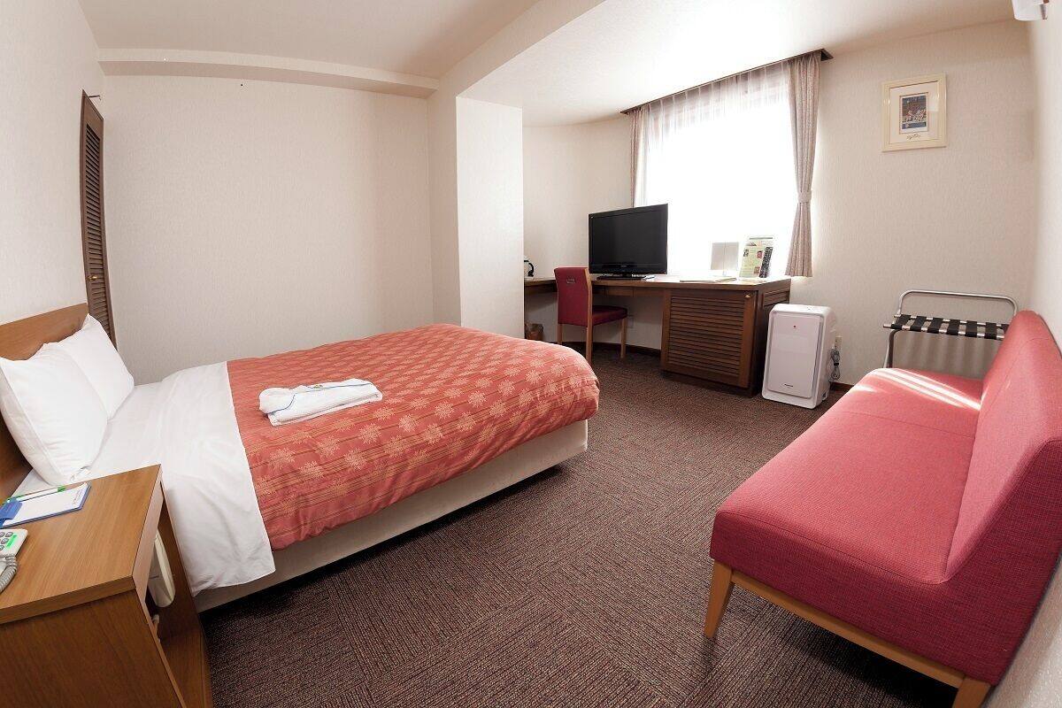 A hotel room with a bed, a desk with a computer, a chair, a TV, and a radiator.