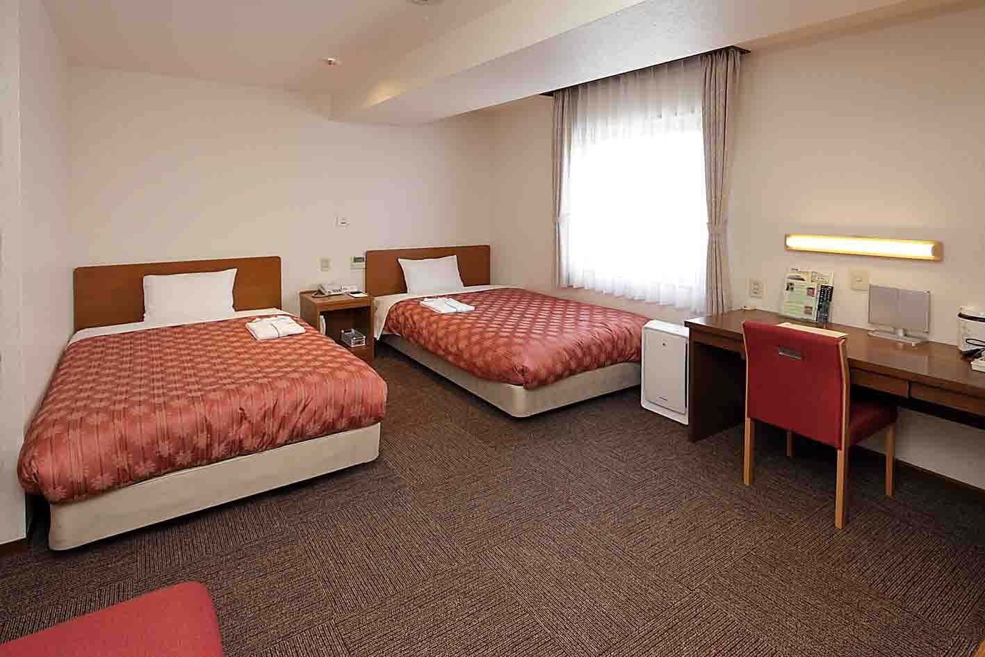 A hotel room with two beds, a desk, a chair, a small table, and a window with curtains.