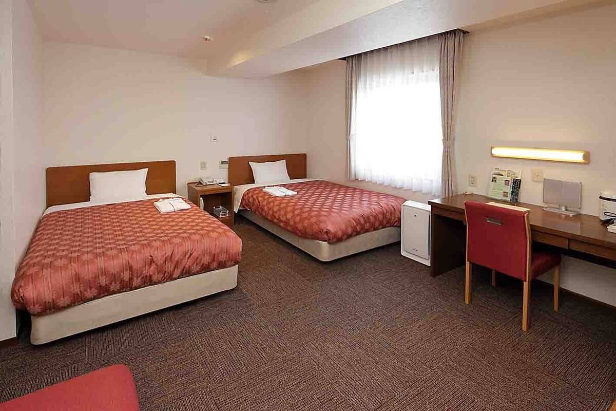 A hotel room with two beds, a desk, a chair, a small table, and a window with curtains.