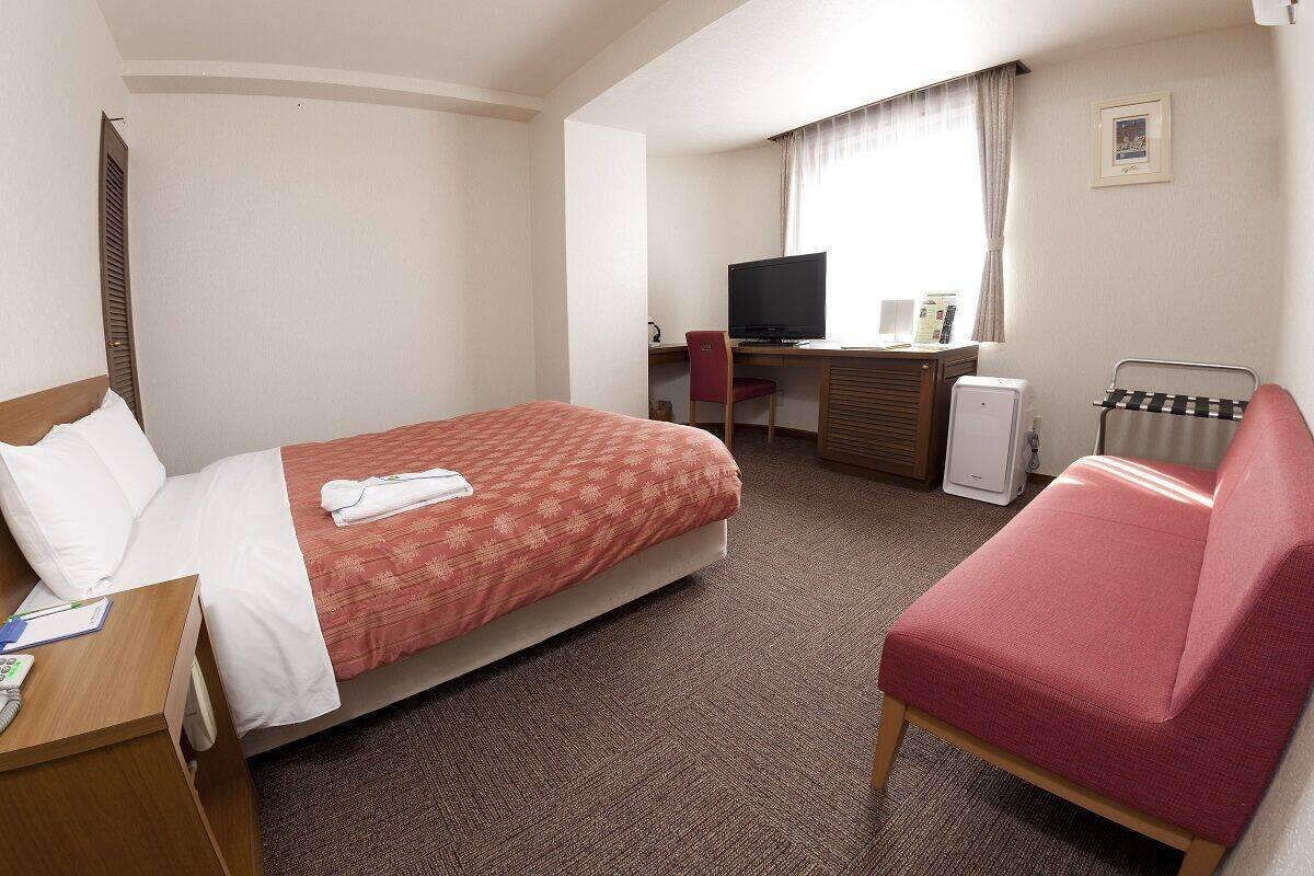 A hotel room with a bed, a desk with a computer, a chair, a radiator, and a TV.