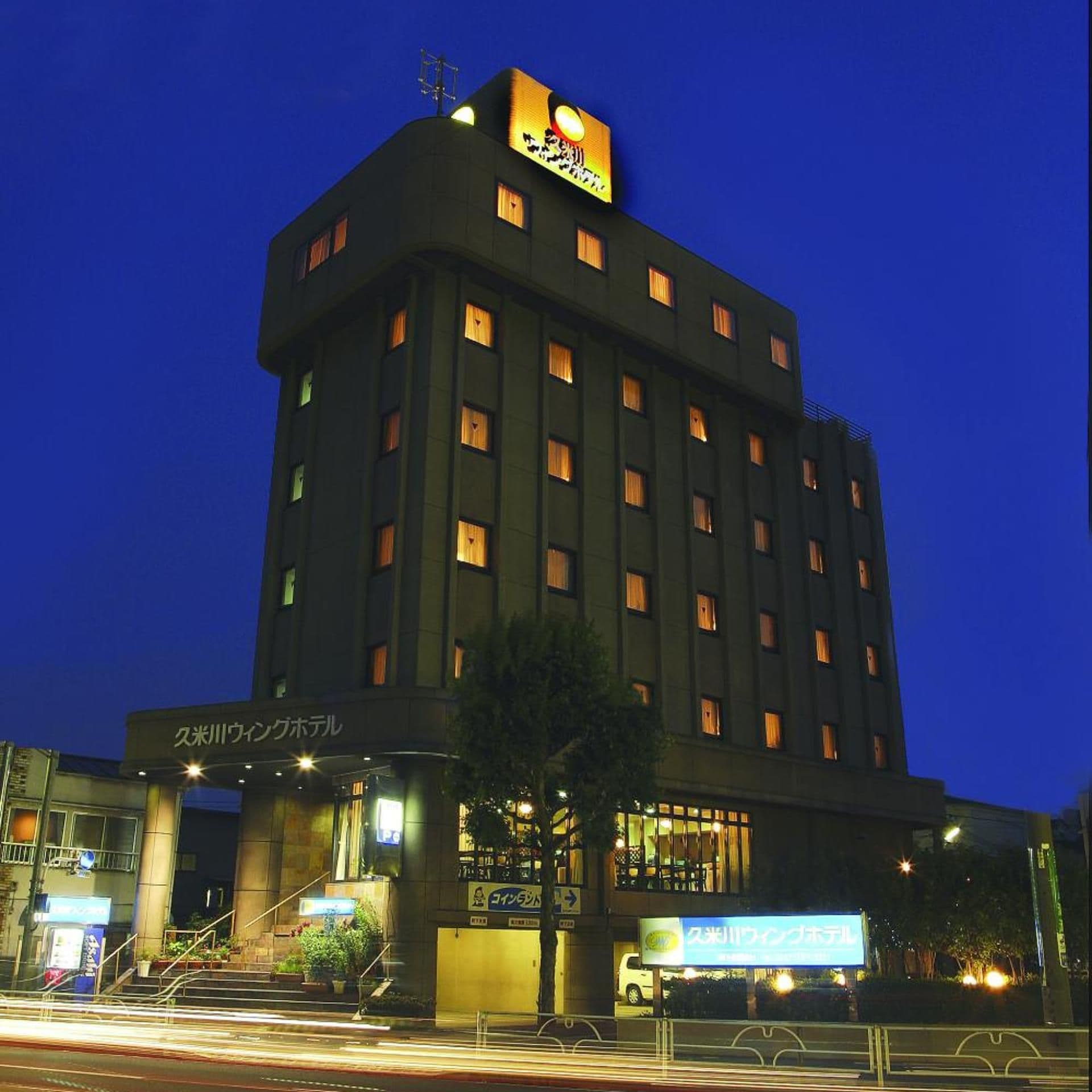 A multi-story hotel with illuminated signage, a visible entrance with steps, and a parking area in front.
