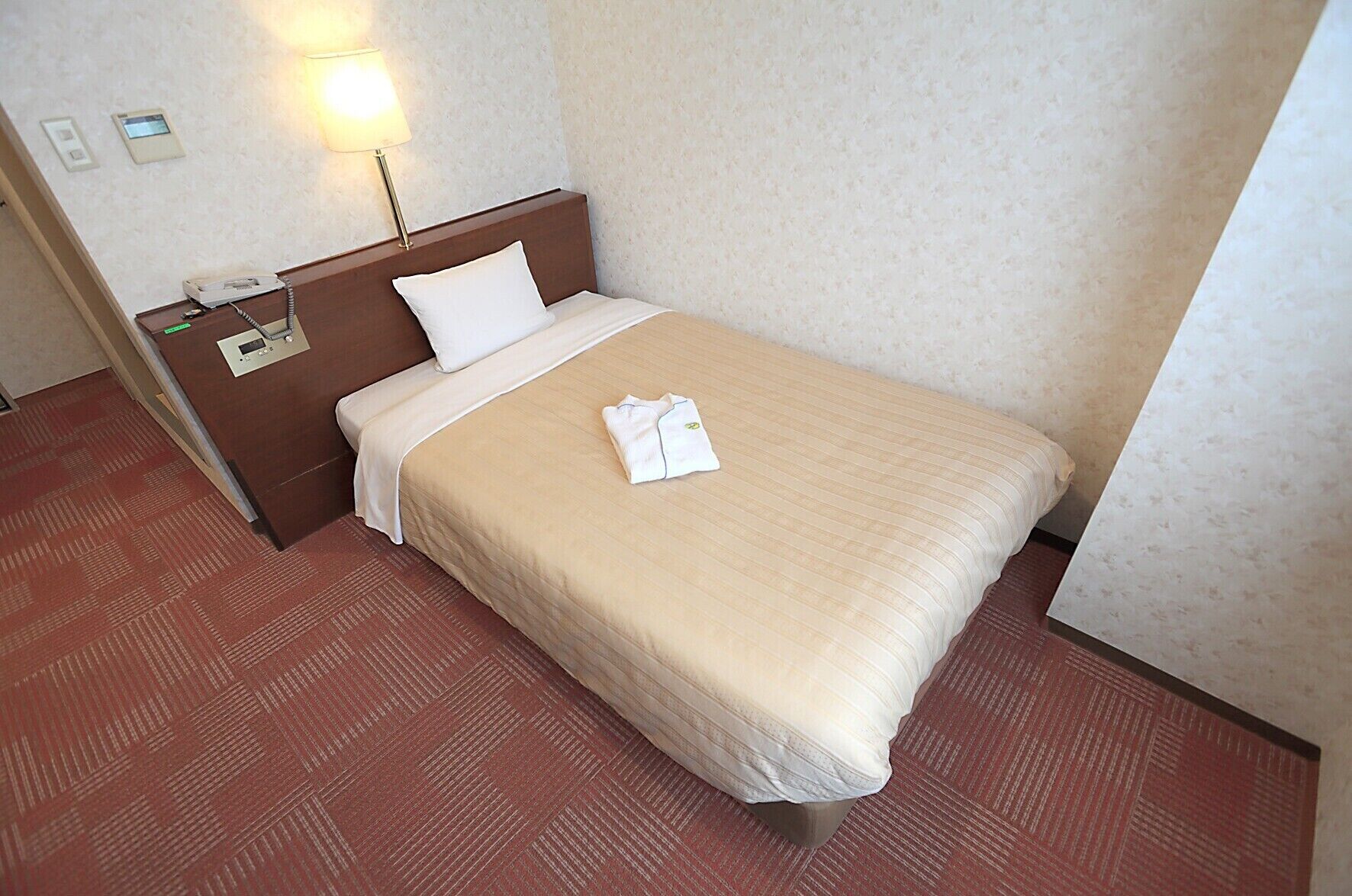 A hotel room with a bed, a nightstand, a lamp, a telephone, and a wall-mounted thermostat.