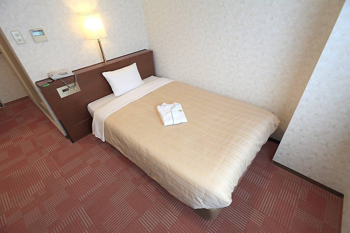 A hotel room with a bed, a nightstand, a lamp, a telephone, and a wall-mounted thermostat.