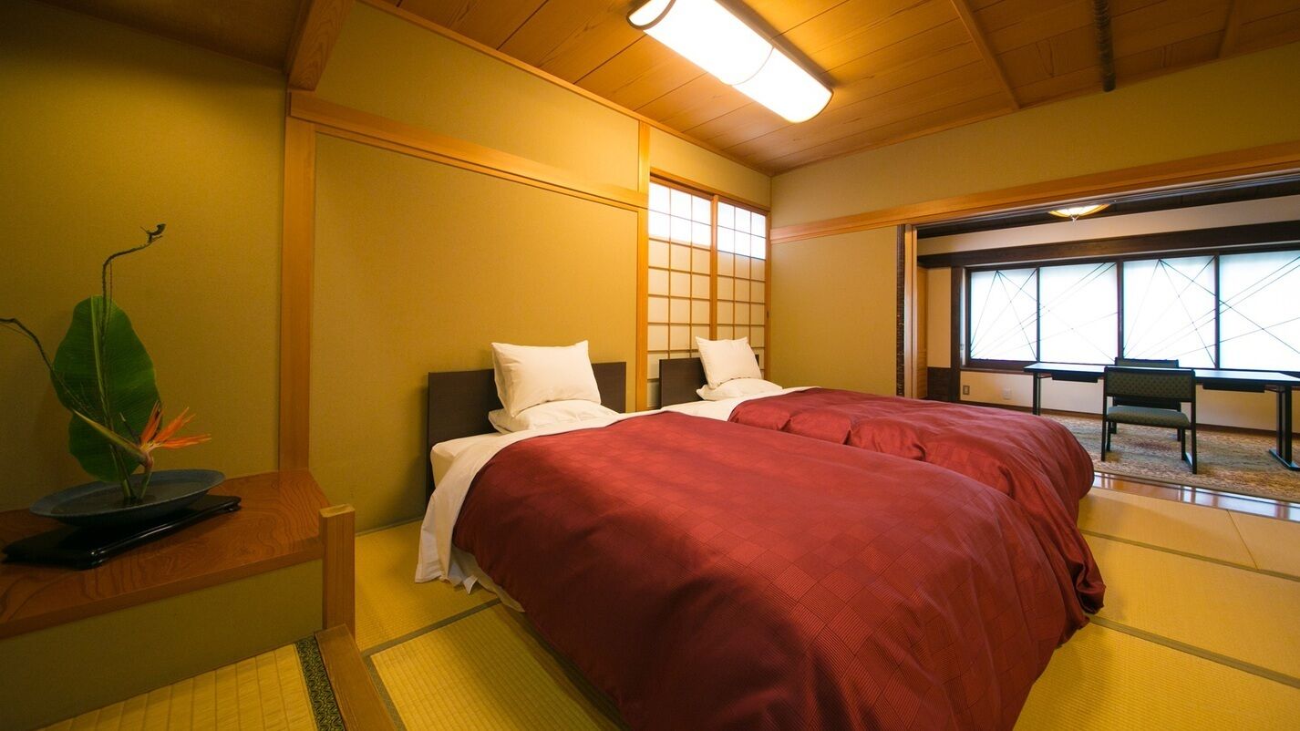 Una habitación con dos camas, cabecera de madera, suelo de esteras de tatami, un ventanal grande y una mesita con una planta.
