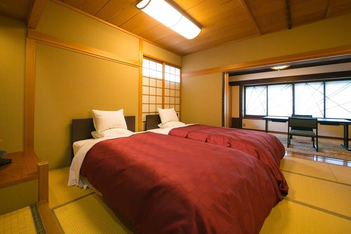 Ein Zimmer mit zwei Betten, einem Holz-Kopfteil, einem Tatami-Bodenbelag, einem großen Fenster und einem kleinen Tisch mit einer Pflanze.
