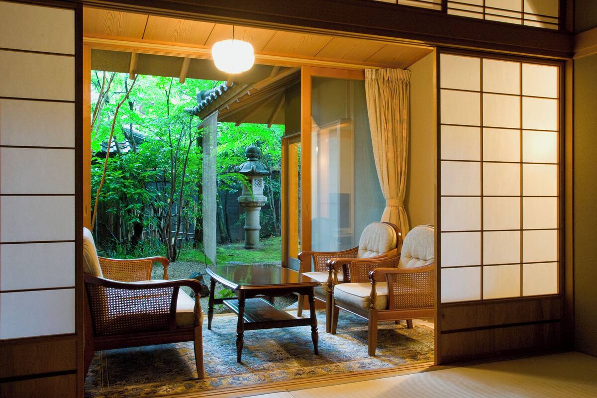 Ein traditioneller japanischer Raum mit Tatami-Böden, einem niedrigen Tisch und zwei Stühlen. Ein großes Fenster gibt den Blick auf einen Garten mit Steinlaterne und viel Grün frei.