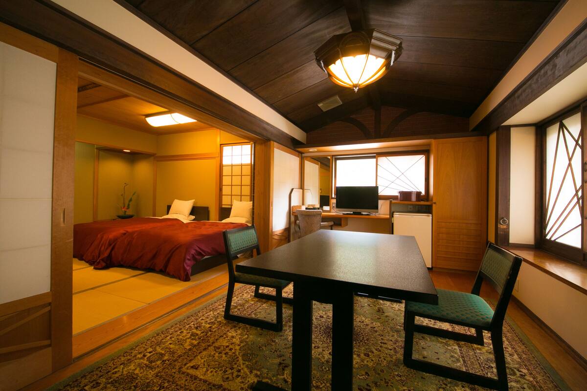 Ein traditioneller japanischer Raum mit Tatami-Böden, einem niedrigen Tisch mit Stühlen, einem Bett mit roter Bettwäsche und einem Fernseher.