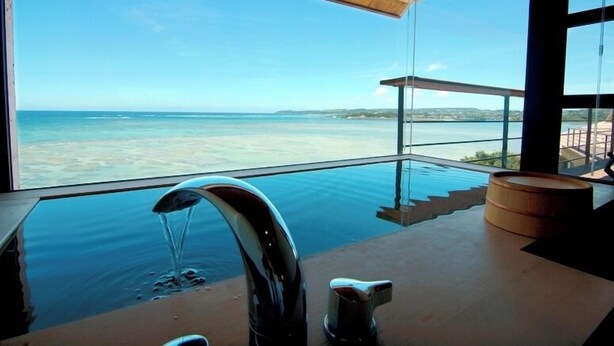 A modern interior with a large glass window overlooking a beach. - Hyakuna Garan (Nanjo)