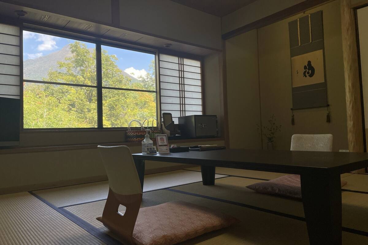 Ein Raum mit Tatami-Matten, einem niedrigen Tisch, einem Sessel, einem Fernseher und Blick auf Bäume durch ein Fenster.