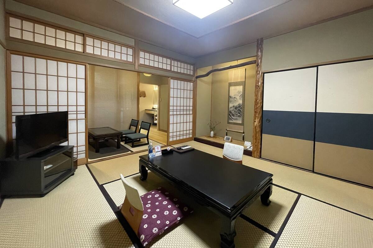 Ein traditioneller japanischer Raum mit Tatami-Boden, einem niedrigen Tisch, einem Fernseher und verschiebbaren Türen.