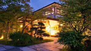 Une maison traditionnelle japonaise, dotée d’une porte d’entrée en bois, entourée d’une végétation luxuriante et de sentiers pavés de pierres.