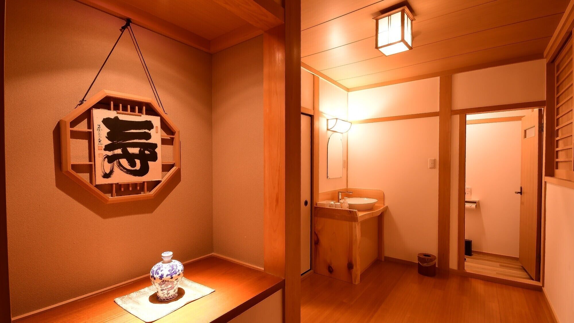 A traditional Japanese interior with a wooden floor, a wall-mounted calligraphy piece, a small table with a decorative item, and a bathroom with a mirror and sink.