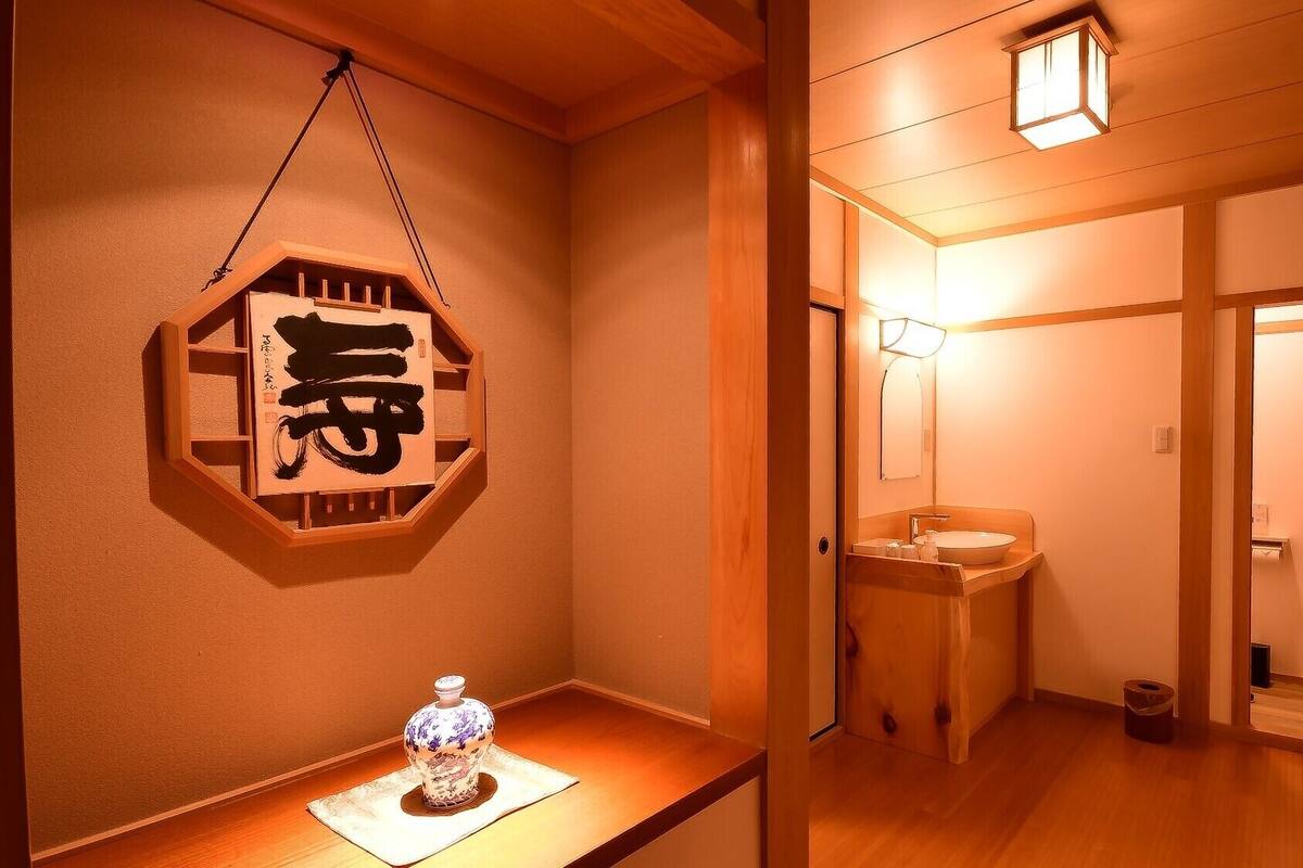 A traditional Japanese interior with a wooden floor, a wall-mounted calligraphy piece, a small table with a decorative item, and a bathroom with a mirror and sink.