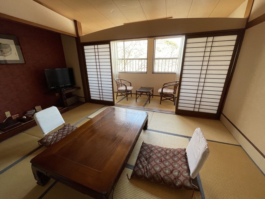 A traditional Japanese room with tatami flooring, a wooden table, two chairs, a television, and sliding shoji doors.