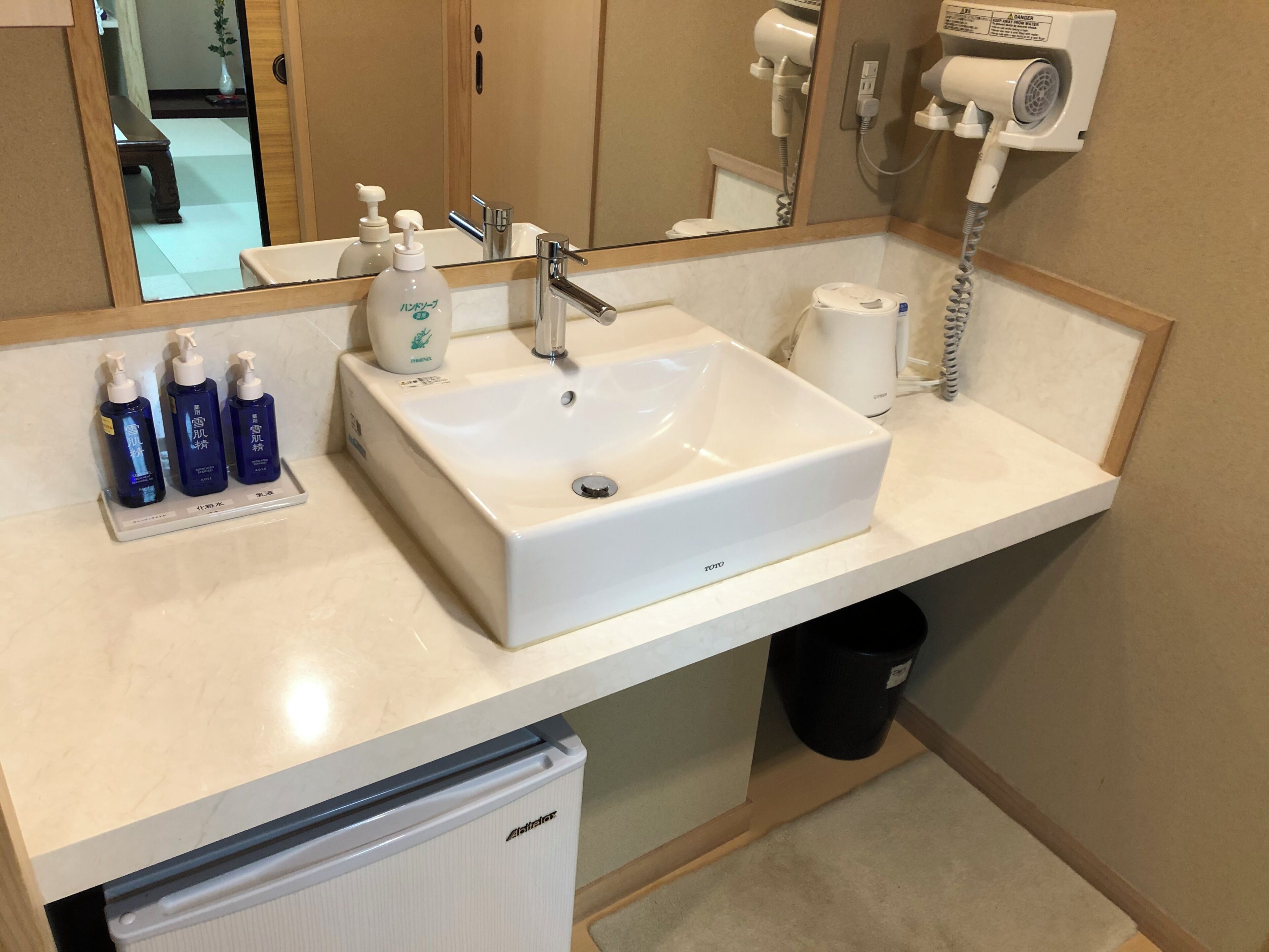 Un baño con un lavabo blanco, un espejo, un secador de pelo y tres botellas de loción.