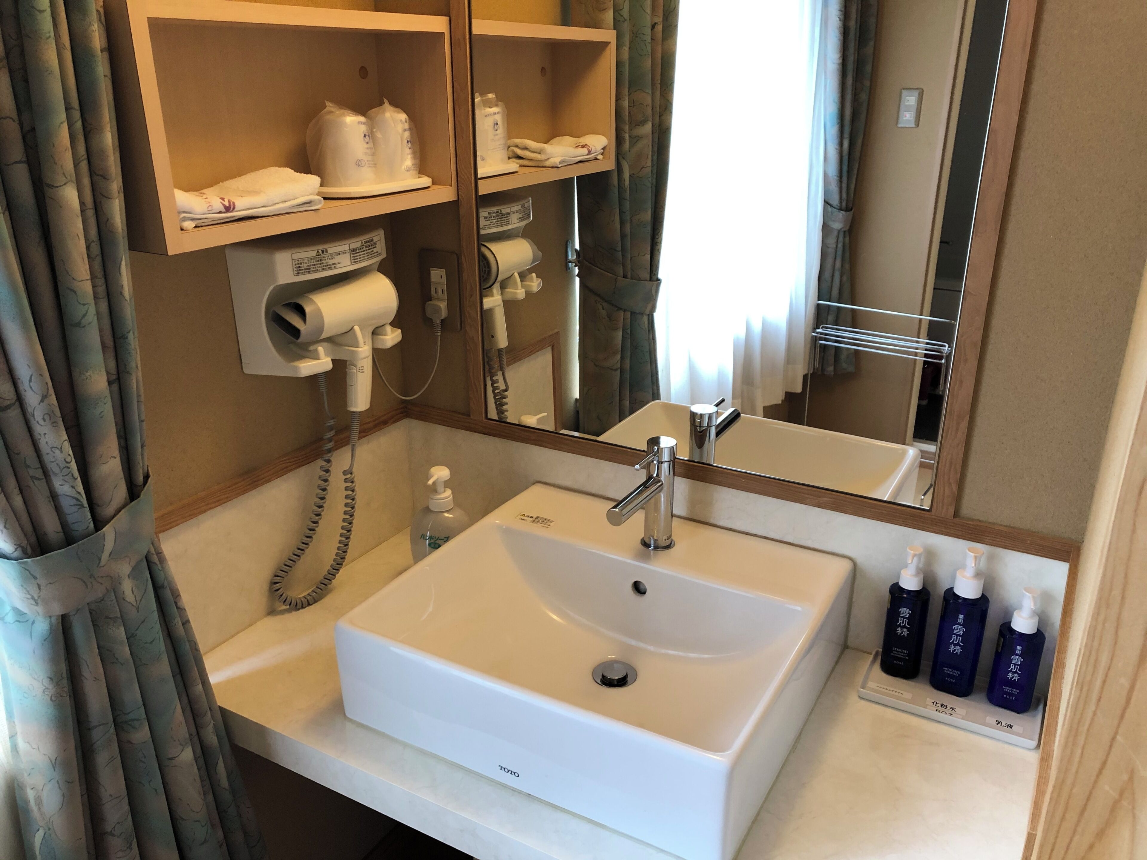 Un baño de hotel con un lavamanos blanco, un espejo, un secador de pelo y artículos de aseo en estantes.