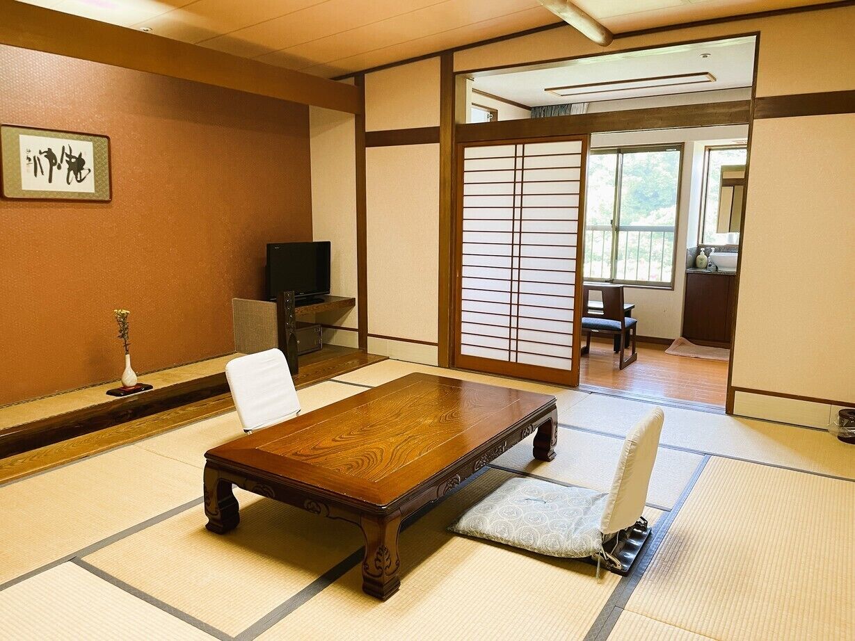 A traditional Japanese room with tatami flooring, a wooden table, a white chair, a television, and a sliding door leading to a balcony.