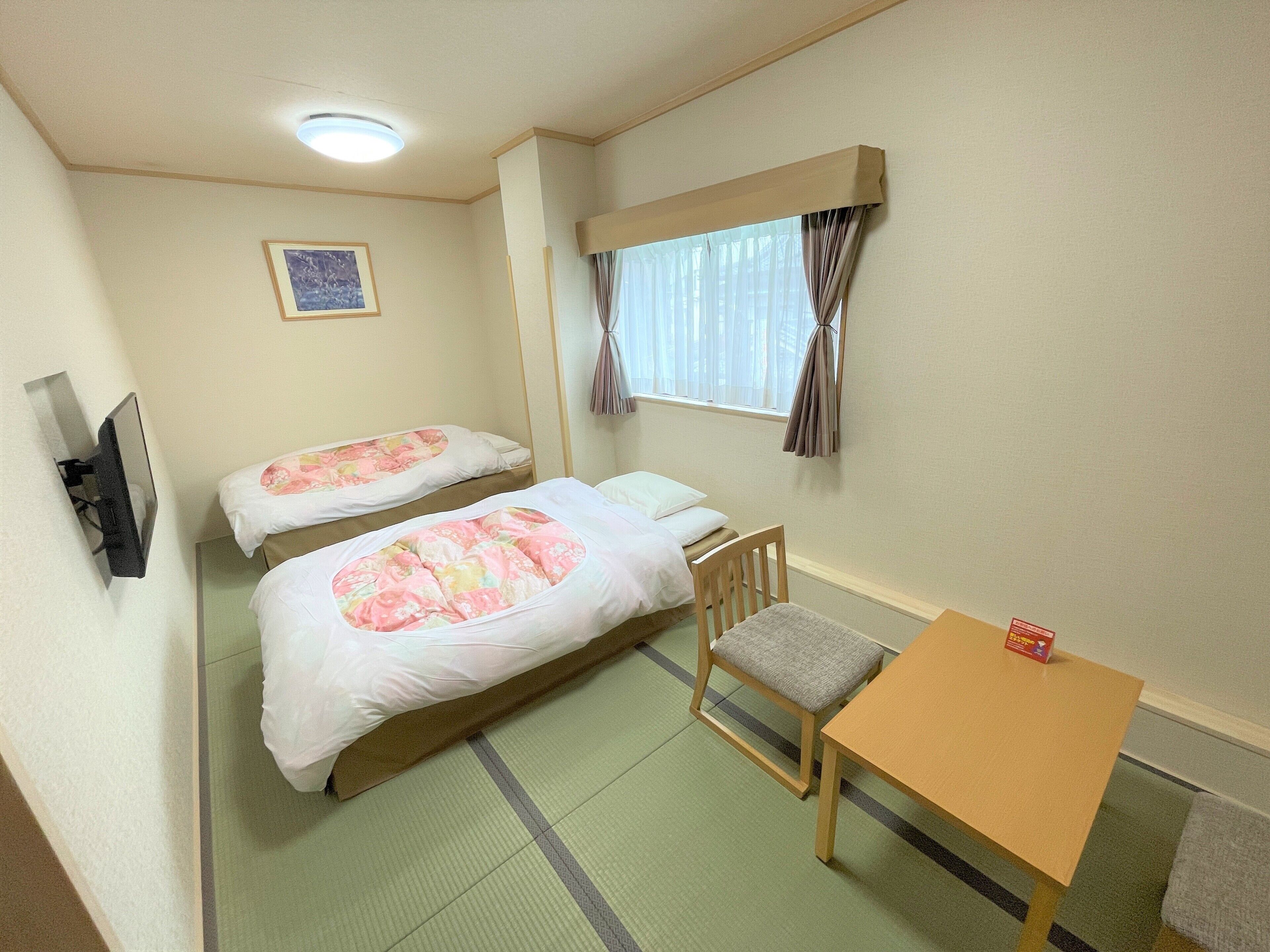 Habitación de hotel con dos camas, una mesita, una silla, un televisor y un ventanal con cortinas.