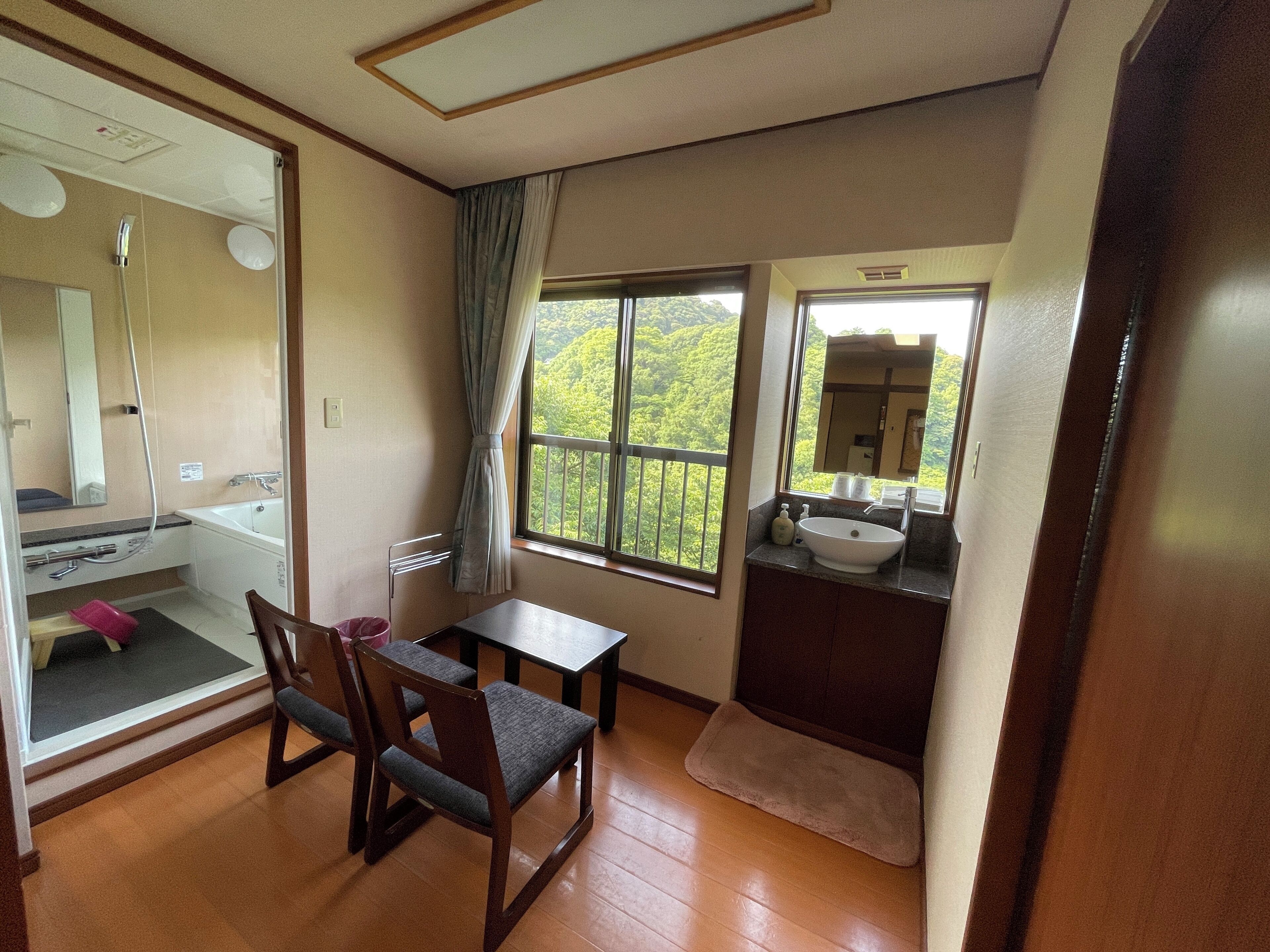Una habitación con piso de madera, una mesa de comedor con sillas, un ventanal con vista a la vegetación, un baño con lavamanos y espejo, y una zona de ducha.