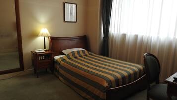 Quarto de hotel com cama de madeira, mesa de cabeceira com abajur, uma cadeira e um espelho grande.