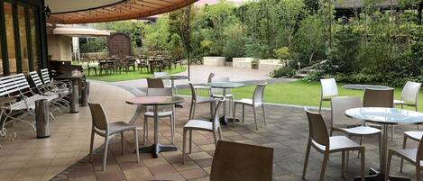 An outdoor patio area with round tables and chairs, a covered seating area, and a view of a garden and building in the background.