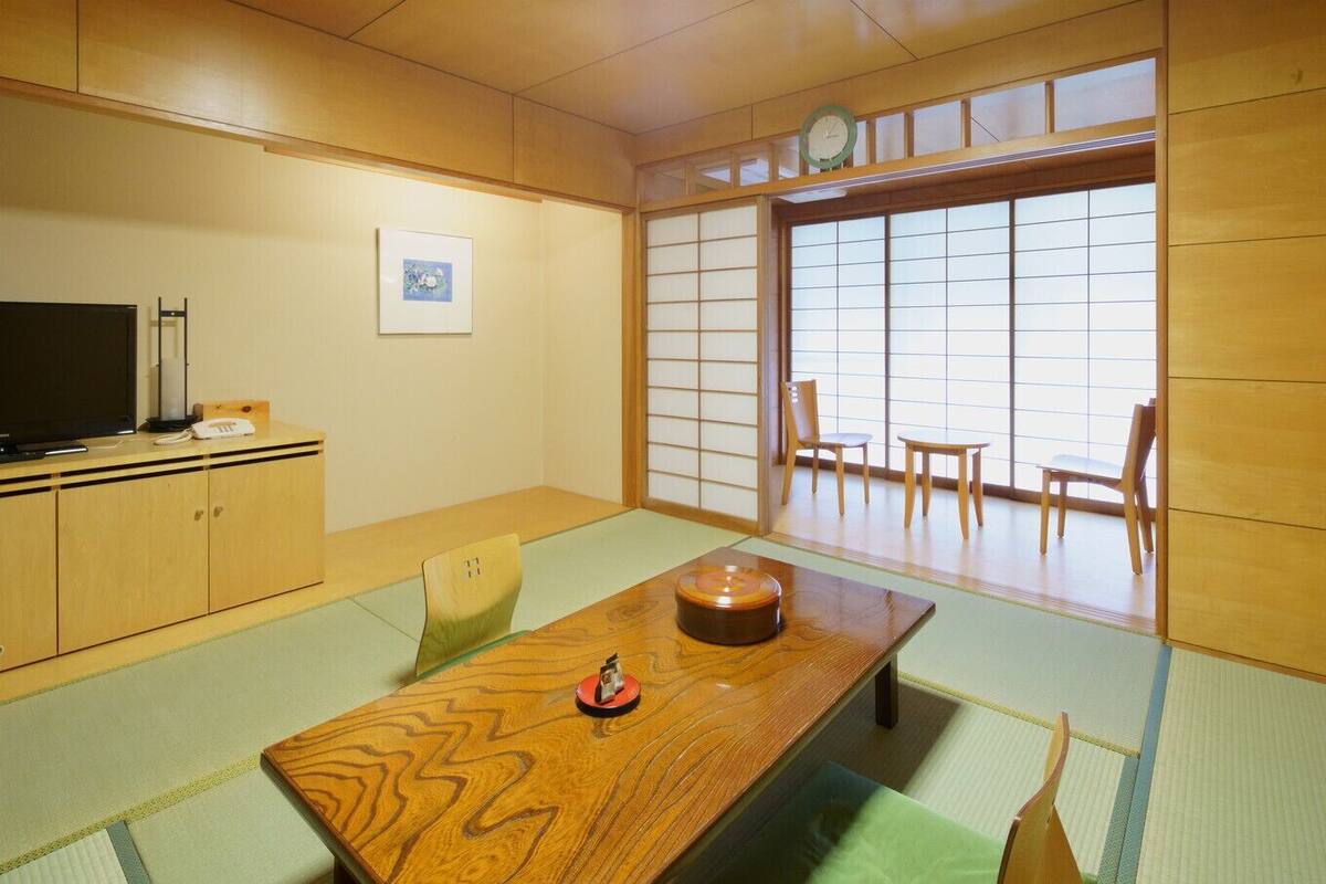 A traditional Japanese room with tatami flooring, a low table with a decorative item, a television on a cabinet, and sliding doors leading to a view.