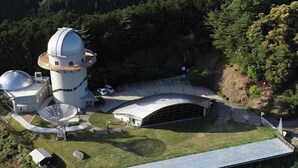 Ett stort observatorium med en central kupol och satellitantenner, omgivet av ett parkliknande område med en böjd byggnad och en utsiktsplattform.