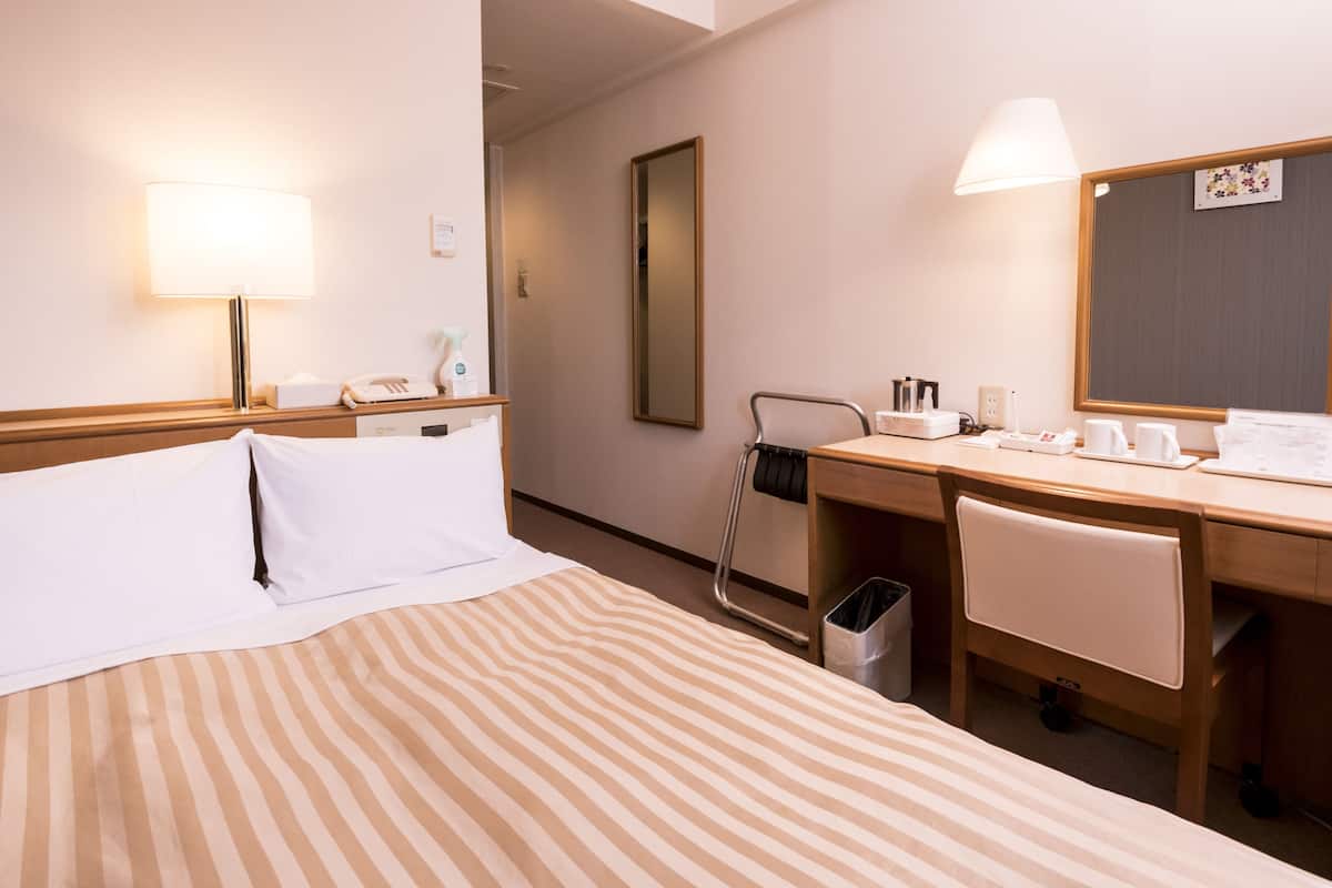 A hotel room with a bed, a desk, a chair, a trash bin, a mirror, and a lamp.
