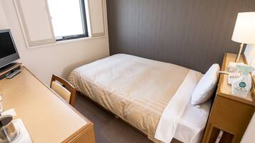 A hotel room with a bed, a desk, a chair, a TV, a lamp, and a nightstand with a bottle and a remote control.