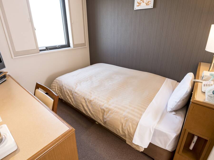 A hotel room with a bed, a desk, a chair, a TV, a lamp, and a nightstand with a bottle and a remote control.